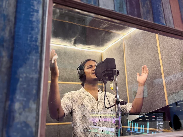 Singer performing in the T Music Lab vocal booth, seen through the control-room glass with the session's level meters reflected